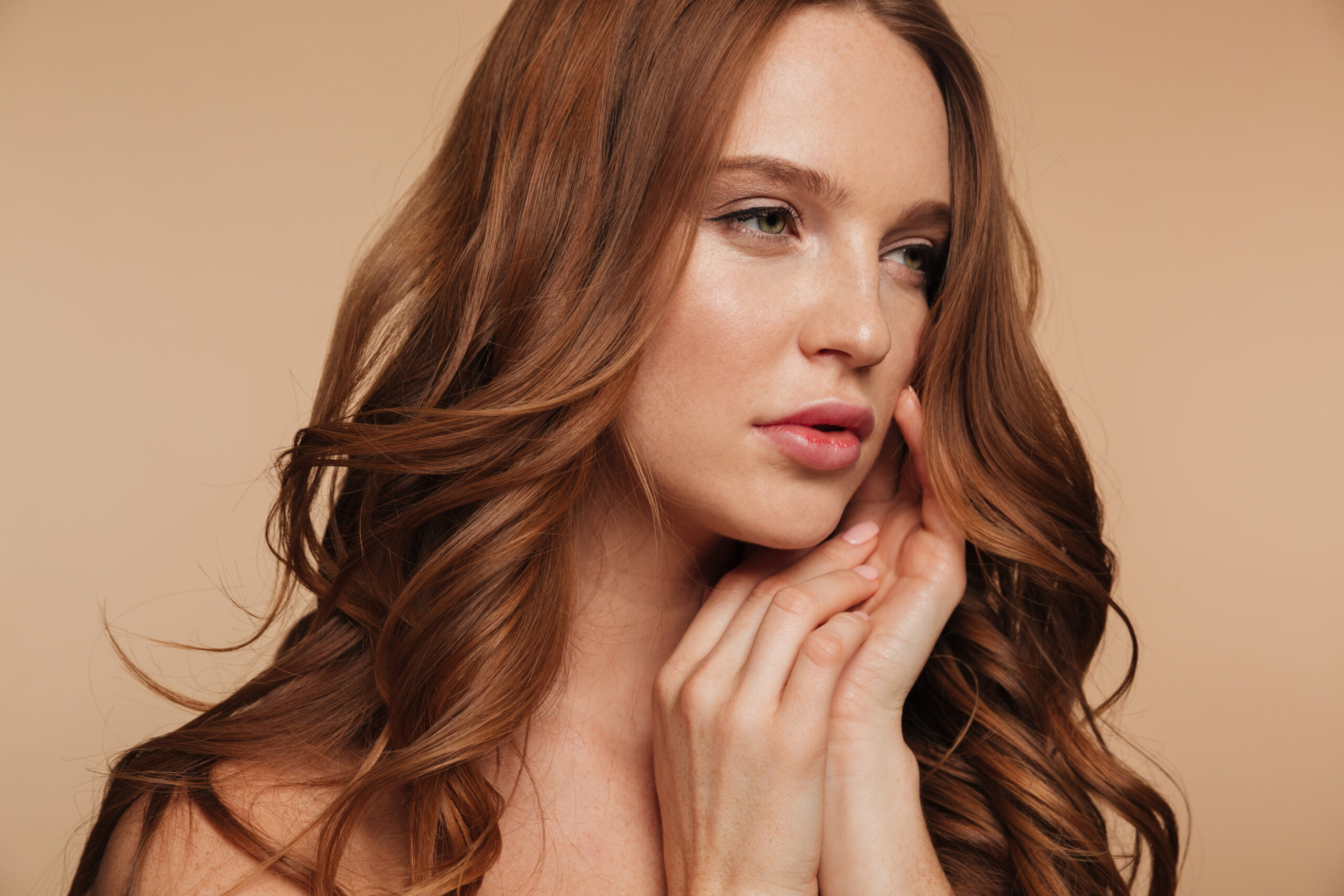 Close up Beauty portrait of pretty ginger woman with long hair looking away while posing with arms near face over cream background